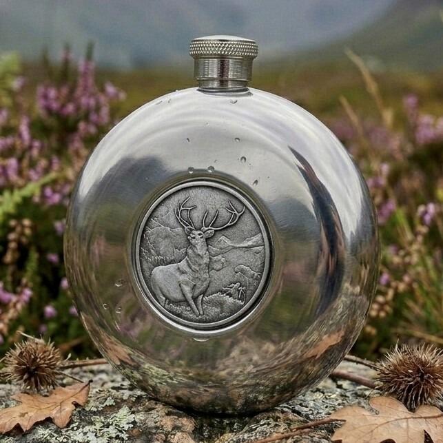 Polished round pewter hip flask featuring a central circular medallion with a detailed embossed stag design, displayed outdoors on a lichen-covered rock with purple heather in the background