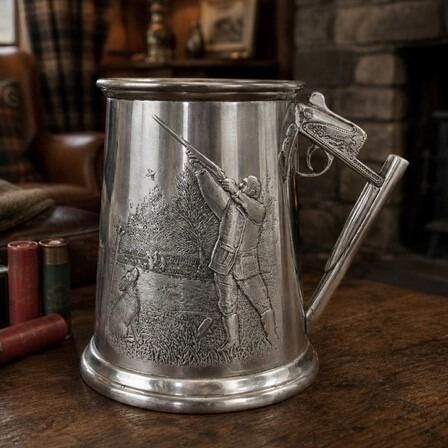 A traditional pewter hunting tankard featuring a detailed embossed relief of a marksman aiming a shotgun at a bird, with a gundog sitting in the grass beside him. The tankard features a unique handle cleverly shaped like the stock, receiver, trigger, and barrels of a shotgun