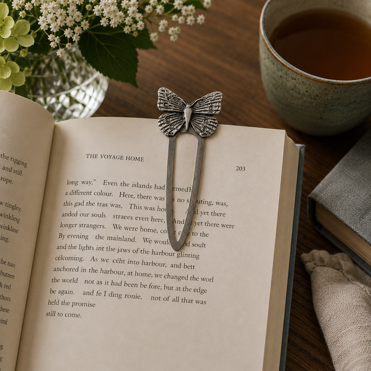 Handcrafted pewter butterfly bookmark fitted over the page of an open book in a cosy reading nook with fresh flowers, a cup of tea and soft natural light