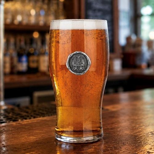 A straight pint beer glass filled with amber ale and a thick white head, featuring a circular pewter wax-seal style badge stamped with a stylized letter A monogram flanked by laurel wreaths