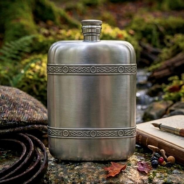 Brushed pewter hip flask featuring two detailed Celtic knotwork bands, styled outdoors on a stone beside a stream with an open vintage journal