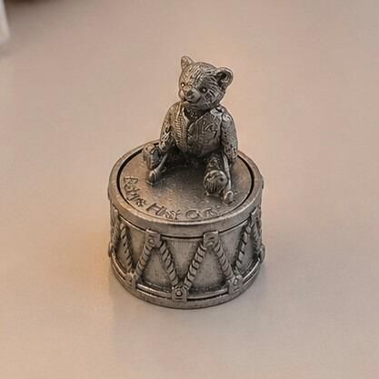 Round pewter keepsake box shaped like a toy drum, engraved with the words "Baby's First Curl" on the lid and topped with a sculpted teddy bear figurine