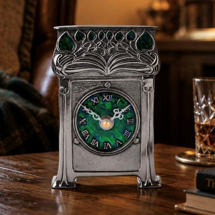 An Arts and Crafts style pewter mantel clock designed by Archibald Knox, featuring a vibrant green enamelled circular face with pewter Roman numerals. The top of the clock is adorned with an ornate, stylized leaf and vine relief design inlaid with green enamel