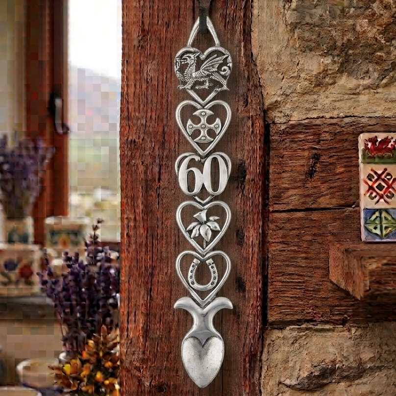Handcrafted pewter 60th birthday Welsh lovespoon hanging on a rustic wooden beam, featuring detailed symbols including a Welsh dragon, Celtic cross, the number 60, a daffodil, a horseshoe, and a heart-shaped spoon bowl.