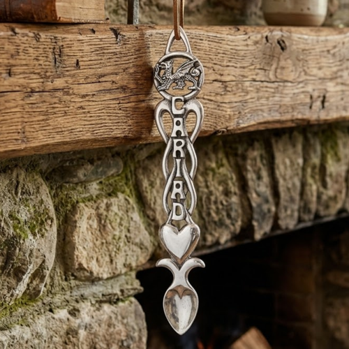 Handcrafted pewter Welsh lovespoon hanging against a wooden beam and stone hearth, featuring a circular Welsh dragon top, the word "CARIAD" carved along open Celtic knotwork, a smooth solid heart, and a spoon bowl.