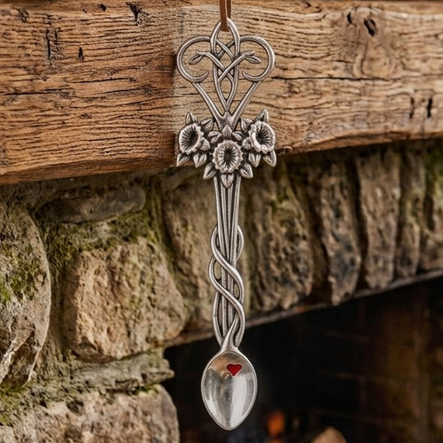 Handcrafted pewter Welsh lovespoon hanging against a wooden beam and stone fireplace, featuring an elegant Celtic knotwork top, a trio of detailed daffodil flowers, a twisted stem, and a red enamel heart accent inside the spoon bowl.