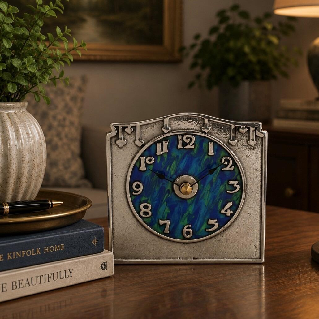 An Arts and Crafts style pewter mantel clock designed by Archibald Knox, featuring a rectangular frame with a gently arched top and a textured finish. The prominent circular clock face has a swirling blue and green enamelled background with raised pewter numerals, accented by small, stylized spade motifs along the upper border.