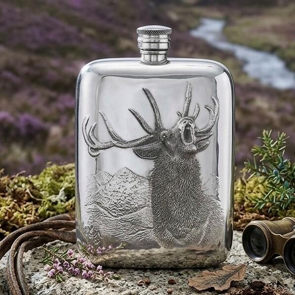 Pewter hip flask engraved with a roaring stag against a mountain backdrop, set outdoors in a Scottish highland heather landscape