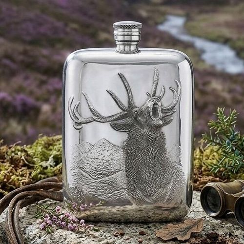 Pewter hip flask engraved with a roaring stag against a mountain backdrop, set outdoors in a Scottish highland heather landscape