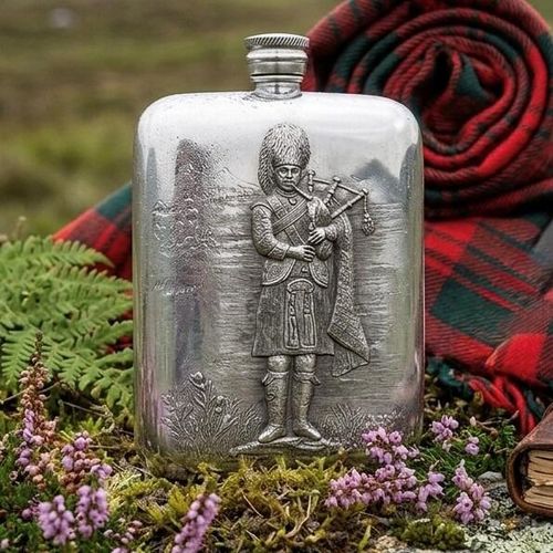Pewter hip flask engraved with a traditional Scottish bagpiper in full kilt attire, set outdoors against a red tartan plaid and highland heather