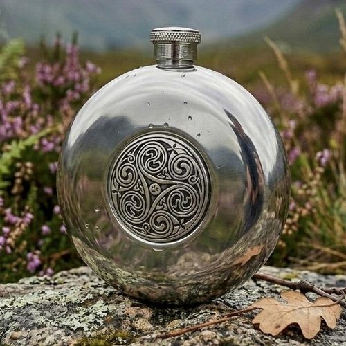 Polished round pewter hip flask featuring a central circular medallion with an intricate Celtic triskele triple spiral design, displayed outdoors on a lichen-covered rock with purple heather in the background