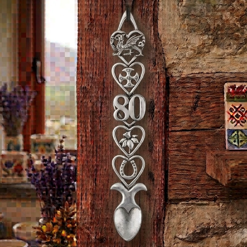 Handcrafted pewter 80th birthday Welsh lovespoon hanging on a rustic wooden beam, featuring detailed symbols including a Welsh dragon, Celtic cross, the number 80, a daffodil, a horseshoe, and a heart-shaped spoon bowl.