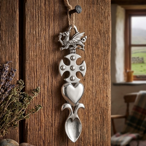 Handcrafted pewter Welsh lovespoon hanging from a twine loop on a rustic wooden beam, featuring a detailed Welsh dragon, a studded Celtic cross, a smooth solid heart, and a textured spoon bowl.
