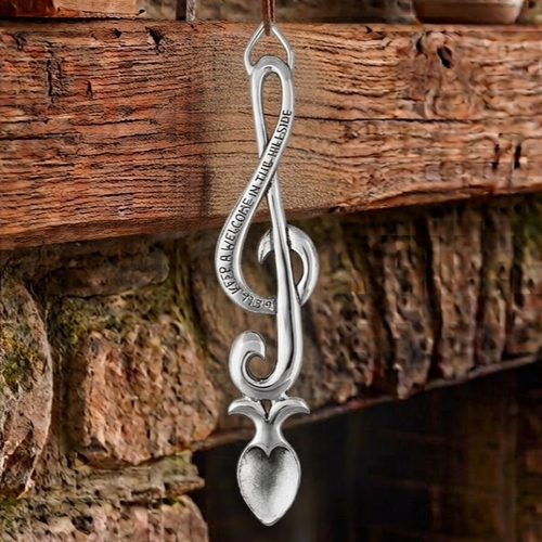Handcrafted pewter Welsh lovespoon hanging against a wooden beam and stone fireplace, shaped like a treble clef engraved with the inscription "WE'LL KEEP A WELCOME IN THE HILLSIDE", leading down to a textured spoon bowl.