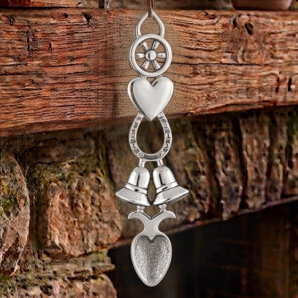 Handcrafted pewter Welsh lovespoon hanging against a wooden beam and stone fireplace, featuring a spoked wheel top, a smooth solid heart, a lucky horseshoe, twin wedding bells, and a textured spoon bowl.