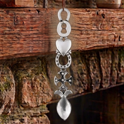 Handcrafted pewter Welsh lovespoon hanging against a wooden beam and stone fireplace, featuring a keyhole lock top, a smooth solid heart, a lucky horseshoe, a studded Celtic cross, and a spoon bowl.