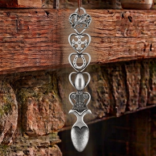 Handcrafted pewter Welsh lovespoon hanging against a wooden beam and stone fireplace, featuring heart-shaped frames enclosing a Welsh dragon, a Celtic cross, a horseshoe, a rugby ball, and the Prince of Wales feathers emblem above a textured spoon bowl.