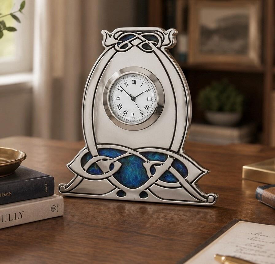 An Arts and Crafts style pewter mantel clock designed by Archibald Knox, featuring a bulbous, horseshoe-shaped frame with an integrated wide base. The clock has a circular white face with black Roman numerals and is accented with intricate Celtic knotwork, including an enamelled top detail and a large, vibrant blue and green enamelled panel stretching across the bottom