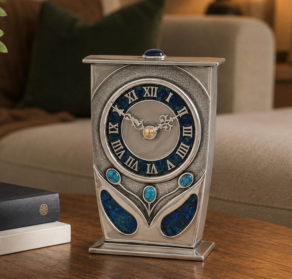An Arts and Crafts style pewter mantel clock designed by Archibald Knox, featuring a tapered rectangular frame with a flat top adorned with a blue cabochon gem. The large circular clock face has a blue and green enamelled chapter ring with silver Roman numerals, while the base is embellished with three turquoise cabochons and two teardrop-shaped panels of blue enamel inlay, resting on a wooden table surface
