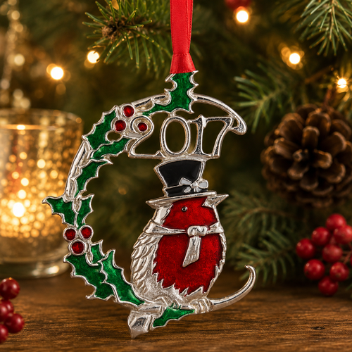 Handcrafted pewter 2017 Christmas ornament featuring an enamelled robin wearing a top hat, surrounded by holly leaves and berries, displayed on a festive Christmas tree with warm holiday lights.