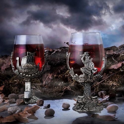 A pair of detailed, Viking-themed wine glasses filled with red wine, each cradled by an intricate pewter wrap and stem. The glass on the left features a pewter relief of a Viking warrior with a shield and axe, resting on a stem shaped like an ornate, knotted Mjölnir (Thor's hammer). The glass on the right features a detailed pewter Viking longship with a sail and carved dragon prow