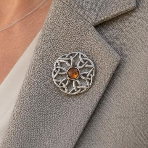 A circular pewter brooch featuring a symmetrical openwork Celtic knot design and a central faceted amber-coloured stone