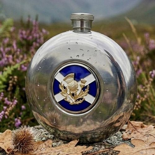 Polished round pewter hip flask featuring a central enameled medallion with the blue and white Scottish Saltire flag topped with a gold-colored Scottish thistle, displayed outdoors on wet stone with heather in the background