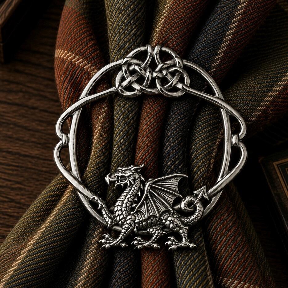 Circular pewter scarf ring featuring a detailed Welsh dragon at the base and a Celtic knotwork top, cinching an earth-toned tartan fabric scarf