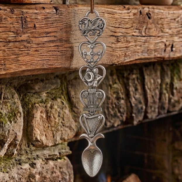 Handcrafted pewter Welsh lovespoon hanging against a wooden beam and stone fireplace, featuring five heart-shaped frames enclosing a Welsh dragon, a Celtic cross, a daffodil, the Prince of Wales feathers emblem, and a harp above a spoon bowl.
