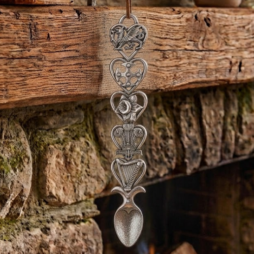 Handcrafted pewter Welsh lovespoon hanging against a wooden beam and stone fireplace, featuring five heart-shaped frames enclosing a Welsh dragon, a Celtic cross, a daffodil, the Prince of Wales feathers emblem, and a harp above a spoon bowl.