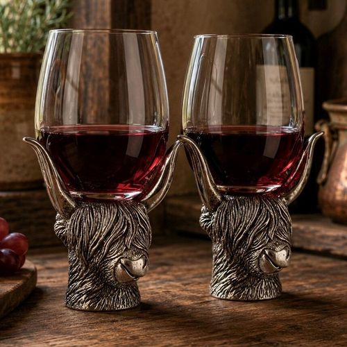 A pair of rustic wine glasses filled with red wine, each supported by a detailed pewter base sculpted in the shape of a Highland cow's head. The pewter stems feature heavily textured, shaggy coats, with the cow's long, sweeping horns curving upward to securely cradle the base of the glass bowls