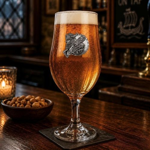 A stemmed beer chalice glass filled with golden ale and a frothy white head, featuring a raised pewter emblem of a detailed Viking warrior profile with a helmet and axe