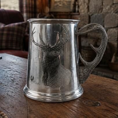 A traditional pewter hunting tankard featuring a detailed embossed relief of a majestic Highland stag with large antlers against a mountain background. The tankard is finished in solid silver-toned pewter and features a unique handle sculpted in the shape of a textured deer antler