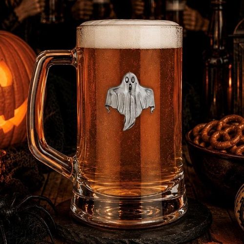 A clear glass beer tankard with a large handle, filled with amber ale and a thick layer of white foam, featuring a detailed pewter emblem of a spooky, flowing ghost with a wide-eyed expression