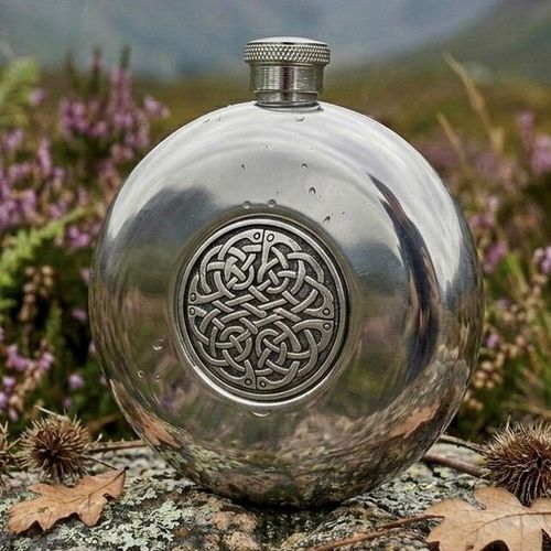 Polished round pewter hip flask featuring a central circular medallion with an intricate Celtic interlace knotwork design, displayed outdoors on a lichen-covered rock with purple heather in the background