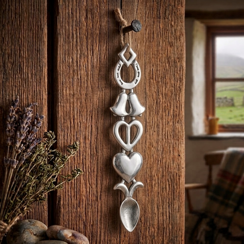 Handcrafted pewter Welsh lovespoon hanging from a twine loop on a rustic wooden beam, featuring a lucky horseshoe, wedding bells, an open heart, a smooth solid heart, and a spoon bowl.