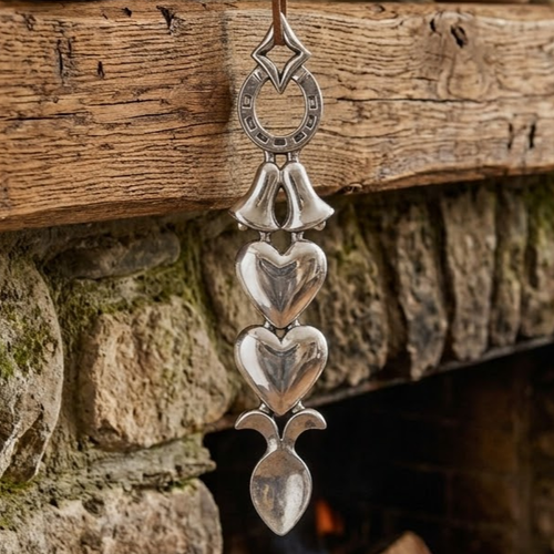 Handcrafted pewter Welsh lovespoon hanging against a wooden beam and stone fireplace, featuring a lucky horseshoe top, twin wedding bells, two smooth solid hearts, and a spoon bowl.