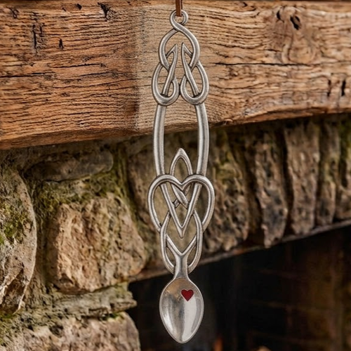 Handcrafted pewter Welsh lovespoon hanging against a wooden beam and stone fireplace, featuring an intricate Celtic knotwork handle, an open central heart motif, and a red enamel heart accent inside the spoon bowl.