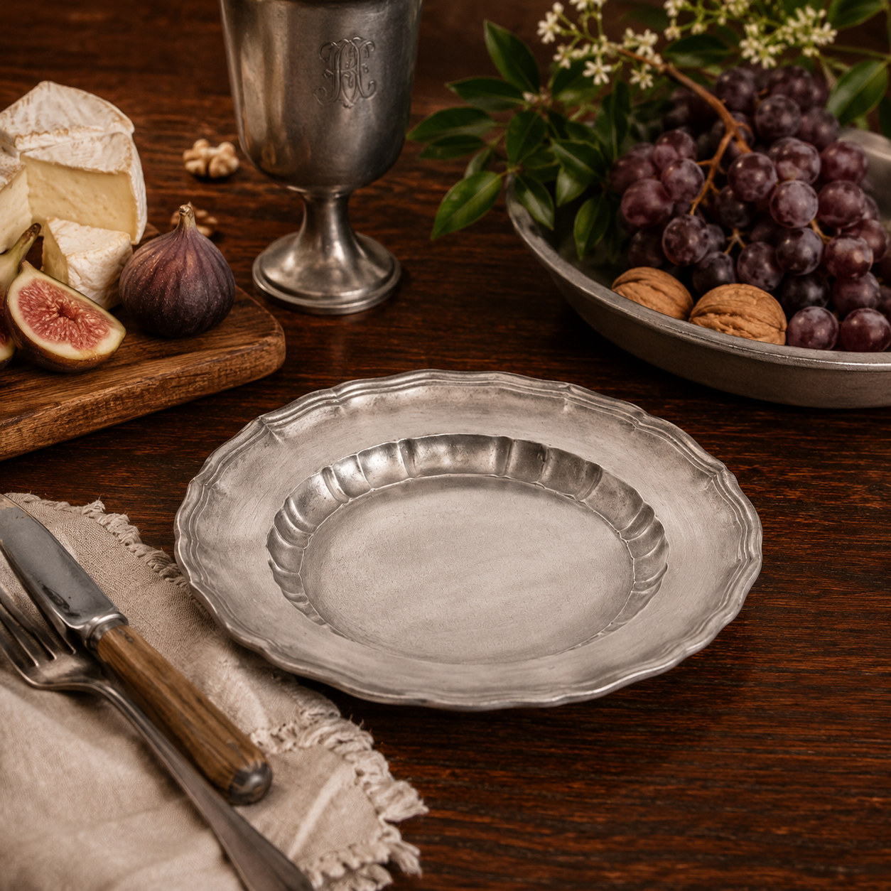 Handcrafted pewter Bavarian scalloped edge plate in antique satin finish displayed on a rustic farmhouse table with artisan bread, figs, grapes, cheese, walnuts and traditional pewter tableware in warm ambient light
