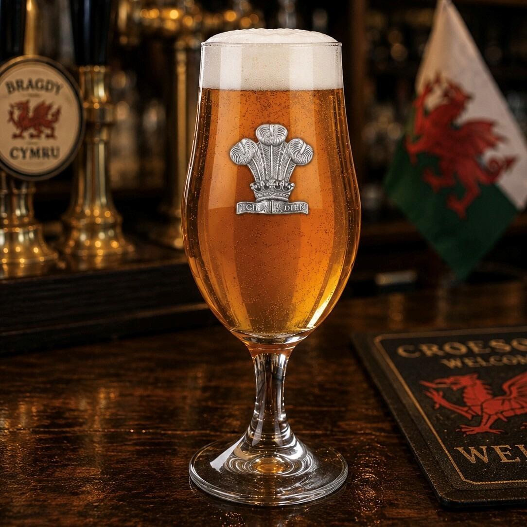 A stemmed beer chalice glass filled with golden ale and a frothy white head, featuring a raised pewter Prince of Wales feathers emblem with detailed plumes and crown