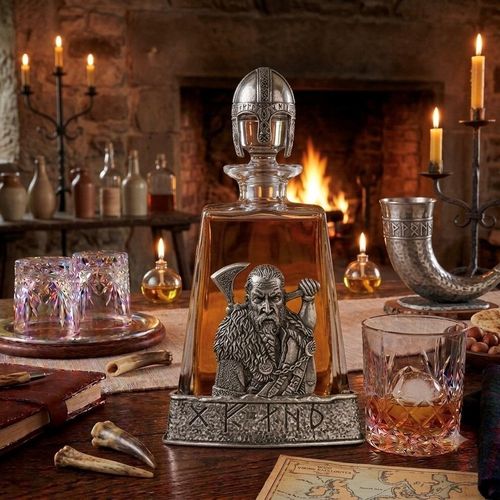 A clear glass spirits decanter filled with amber liquid, sitting in a hammered-effect pewter base engraved with Nordic runes. The front of the decanter is adorned with a detailed pewter relief of a bearded Viking warrior with a braided beard, wearing a fur cloak and holding a battle axe over his shoulder. The bottle is sealed with a glass stopper topped by a sculpted pewter Viking helmet