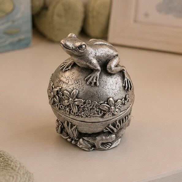Spherical pewter tooth fairy keepsake box featuring a sculpted frog figurine sitting on a dome lid decorated with a relief of flowers, displayed on a table with nursery elements