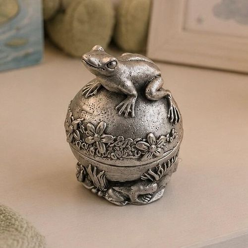 Spherical pewter tooth fairy keepsake box featuring a sculpted frog figurine sitting on a dome lid decorated with a relief of flowers, displayed on a table with nursery elements