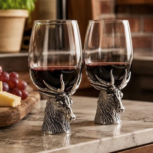 A pair of rustic wine glasses filled with red wine, each supported by a detailed pewter base sculpted in the shape of a stag's head. The pewter stems feature a textured, fur-like finish on the deer's neck, with the majestic, multi-pointed antlers sweeping upward to securely cradle the sides of the glass bowls