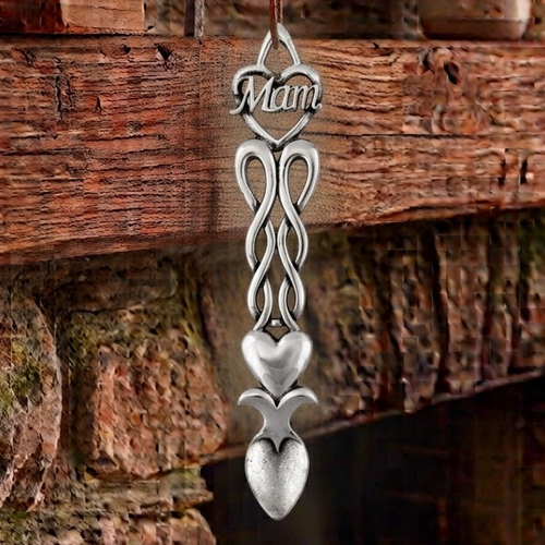 Handcrafted pewter Welsh lovespoon hanging against a wooden beam and stone fireplace, featuring a heart top with script lettering reading "Mam", flowing ribbon knotwork, a smooth solid heart center, and a spoon bowl.