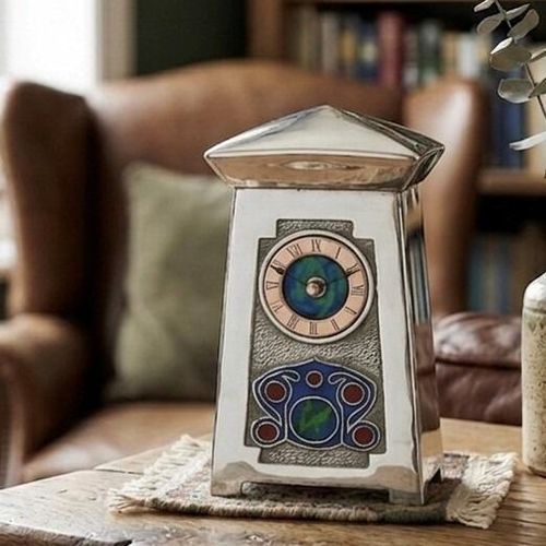 An Arts and Crafts style pewter mantel clock designed by Archibald Knox, featuring a tapered body topped with a distinct, overhanging gabled or pyramidal roof. The circular clock face has a copper chapter ring with Roman numerals surrounding a blue and green enamelled centre, set into a textured rectangular front panel that also contains a colourful Celtic-inspired enamel motif at the base