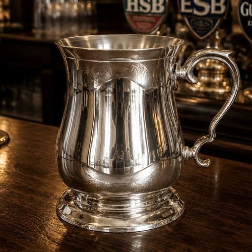 A traditional Georgian-style pewter tankard with a highly polished, reflective finish and a classic tulip-shaped bell body. The tankard features a wide flared rim, a stepped circular base, and an elegantly curved scroll handle. It sits on a dark wooden bar counter in a pub setting