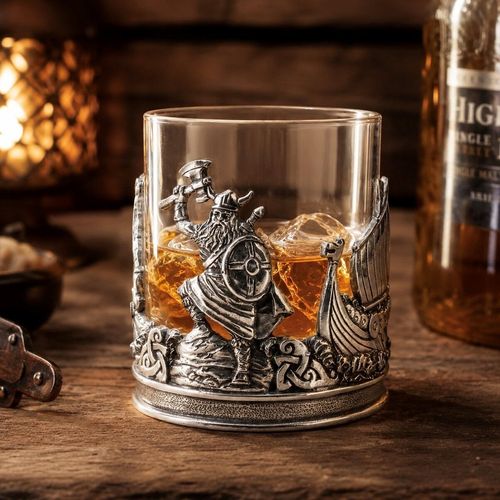 A lowball glass whisky tumbler filled with amber spirits and ice cubes, encased in an ornate, sculpted pewter holder. The front of the holder features a detailed relief of a bearded Viking warrior in a horned helmet, raising a battle axe and holding a round shield. To his side, a carved Viking longship cuts through waves, all resting above a textured base decorated with continuous Celtic knotwork bands