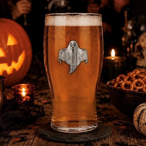 A clear pint beer glass filled with golden amber ale and a frothy white head, featuring a detailed pewter ghost motif in the center