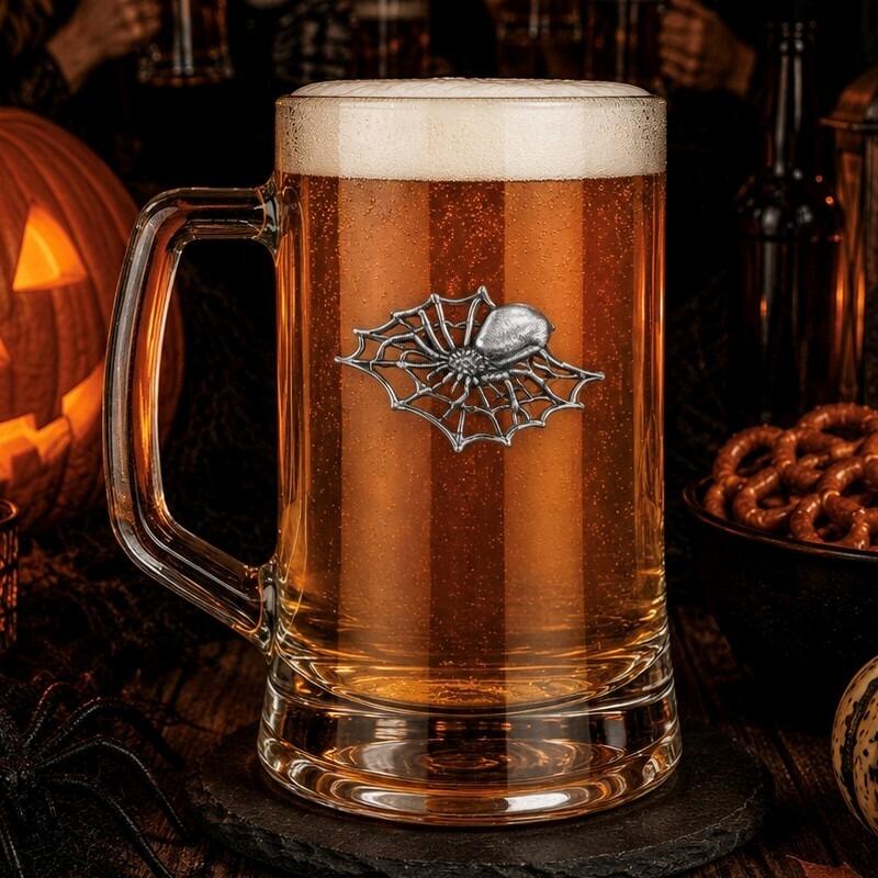 A clear glass beer tankard with a sturdy handle, filled with amber lager and a thick white foam head, featuring a detailed pewter emblem of a spider sitting on an intricate spiderweb